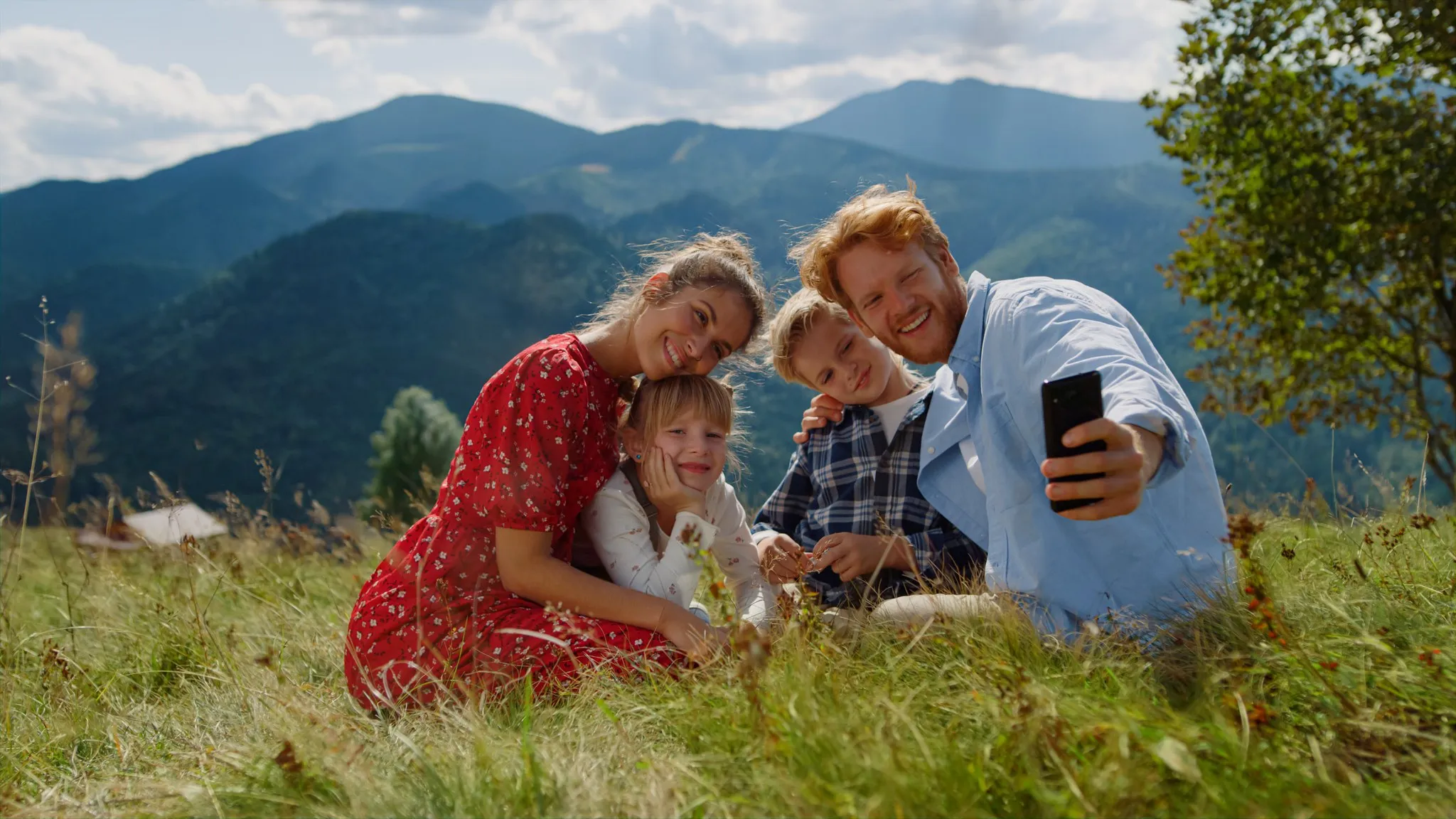 Familie mit zwei Kindern sitzt auf einer Bergwiese und macht ein Selfie, im Hintergrund bewaldete Bergketten