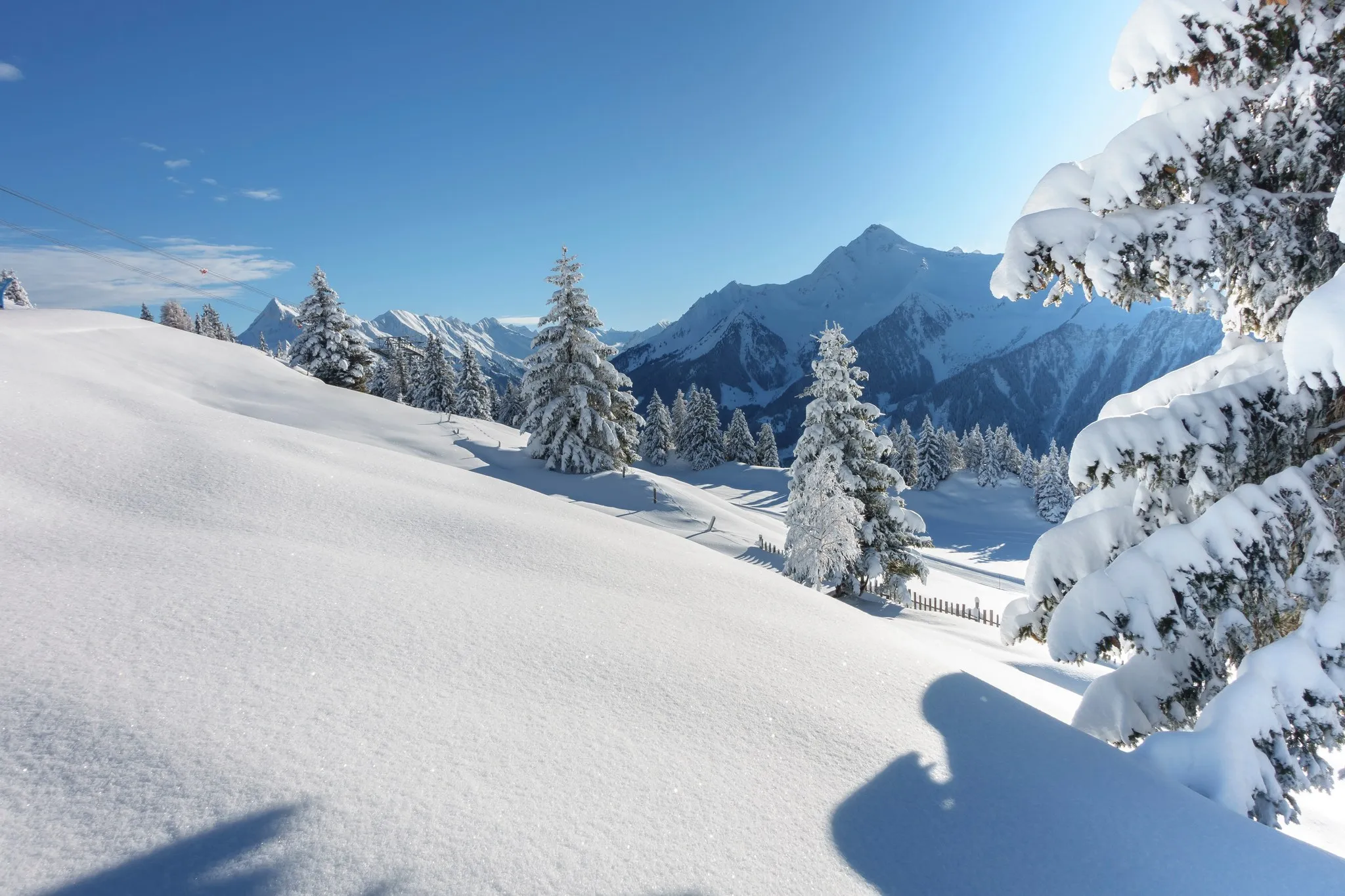 Tief verschneiter Hang mit schneebedeckten Fichten, dahinter eine sonnenbeschienene Bergkette unter blauem Himmel