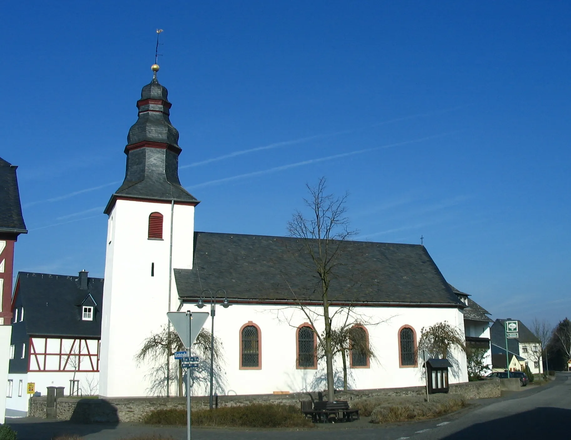 Weiß verputzte Dorfkirche mit schiefergedeckter Haube, daneben ein Fachwerkhaus, kahle Bäume vor blauem Winterhimmel