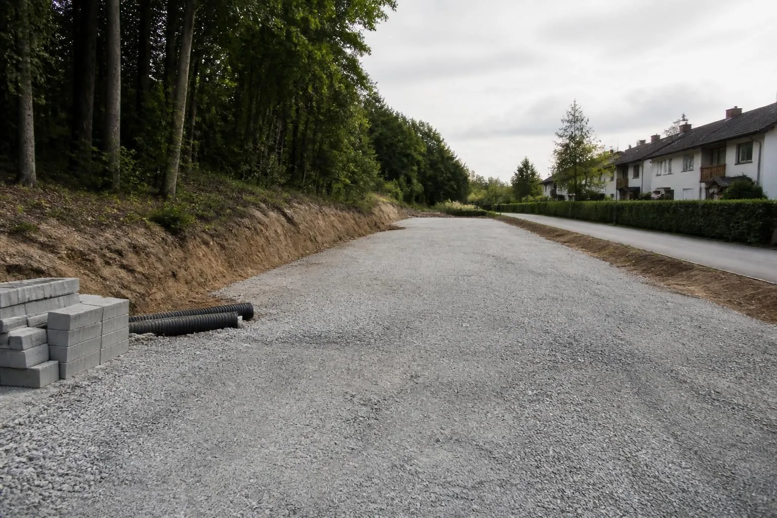Frisch geschotterte Fläche eines Parkplatzes im Bau, links ein bewaldeter Hang, rechts eine Wohnstraße, Bordsteine gestapelt