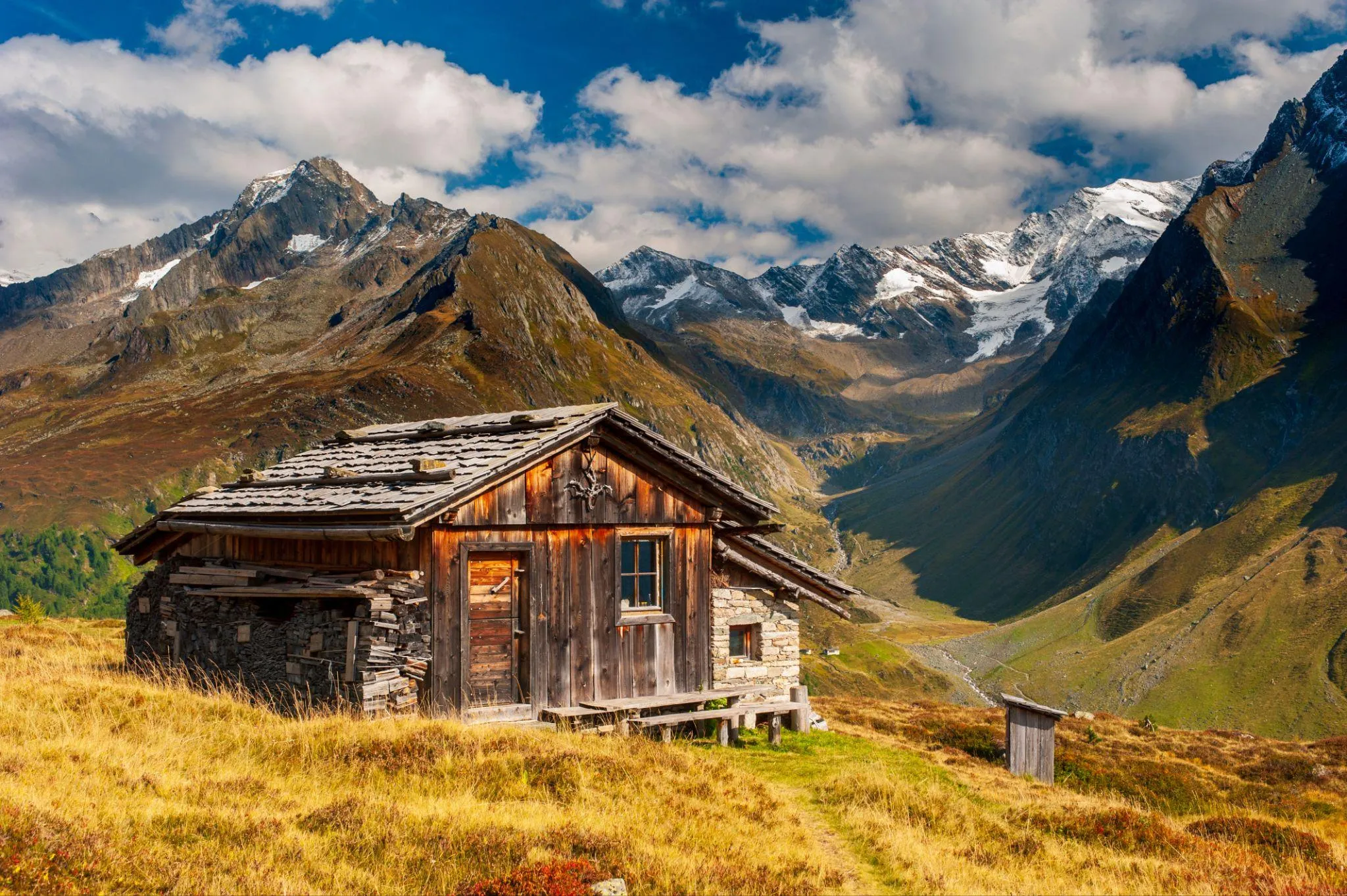 Almhütte aus Holz und Bruchstein auf einer herbstlichen Bergwiese, dahinter ein Talkessel mit schneebedeckten Gipfeln