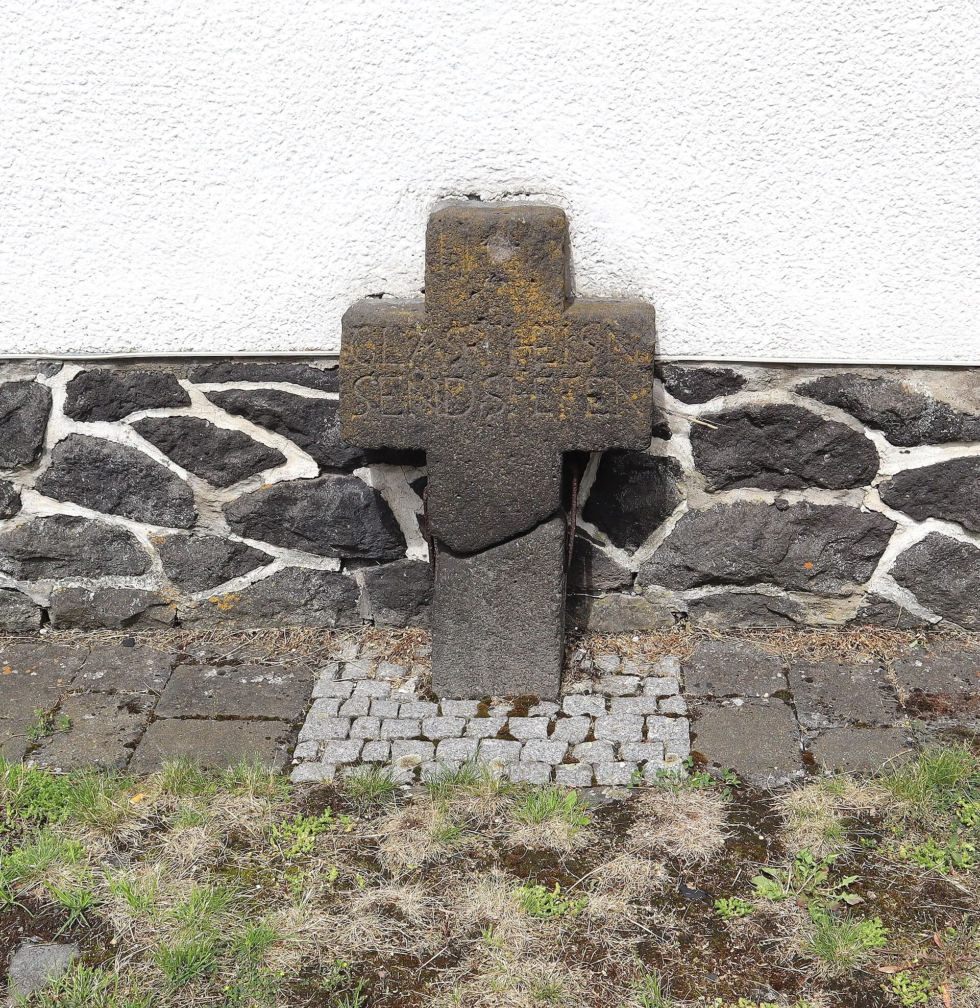 Verwittertes Steinkreuz vor einer dunklen Bruchsteinmauer an der Kapelle St. Bartholomäus in Hauroth
