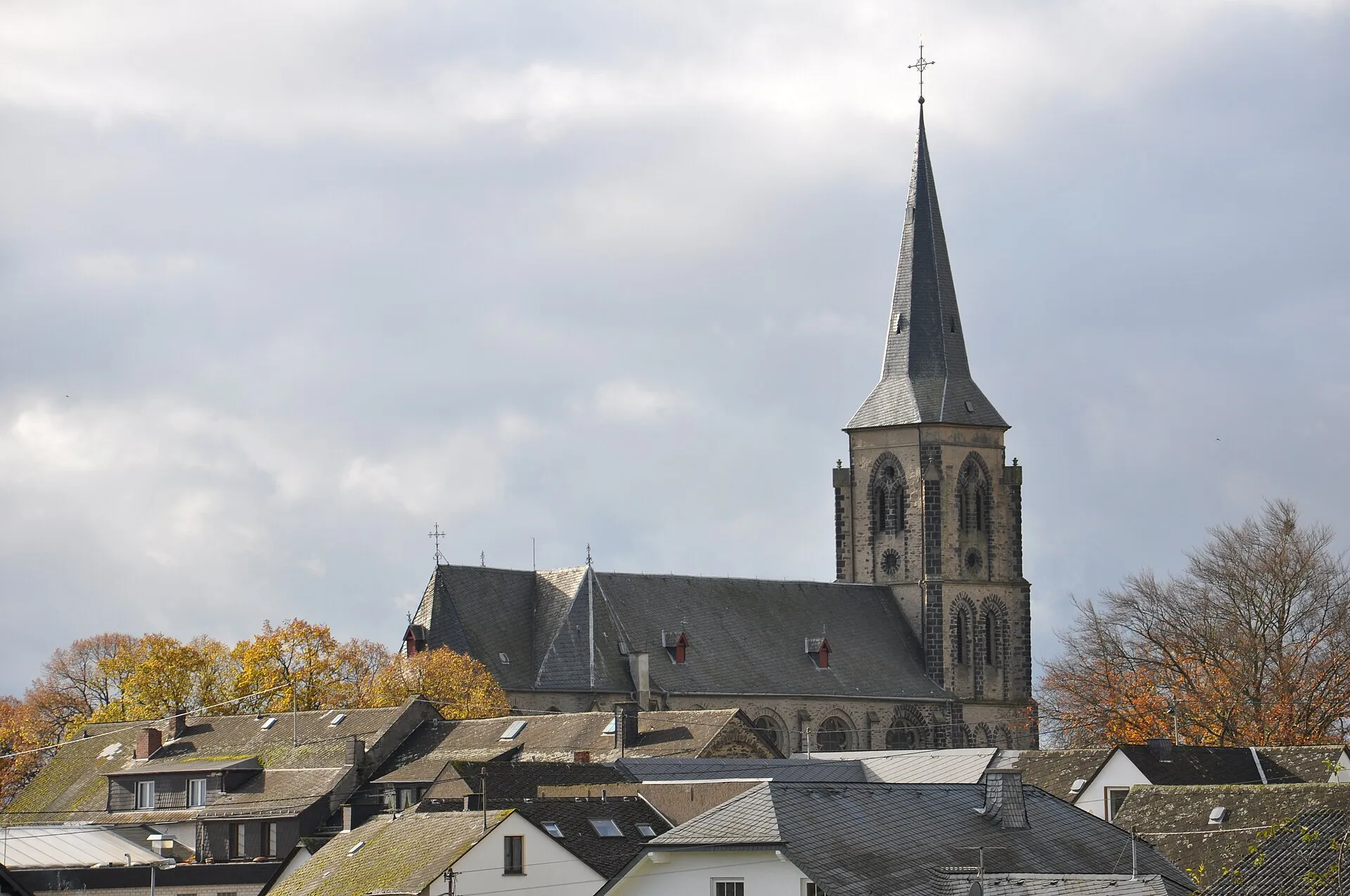 Neugotische Kirche mit hohem Schieferturm über den Dächern eines Eifeldorfes, davor Herbstbäume unter grauem Himmel