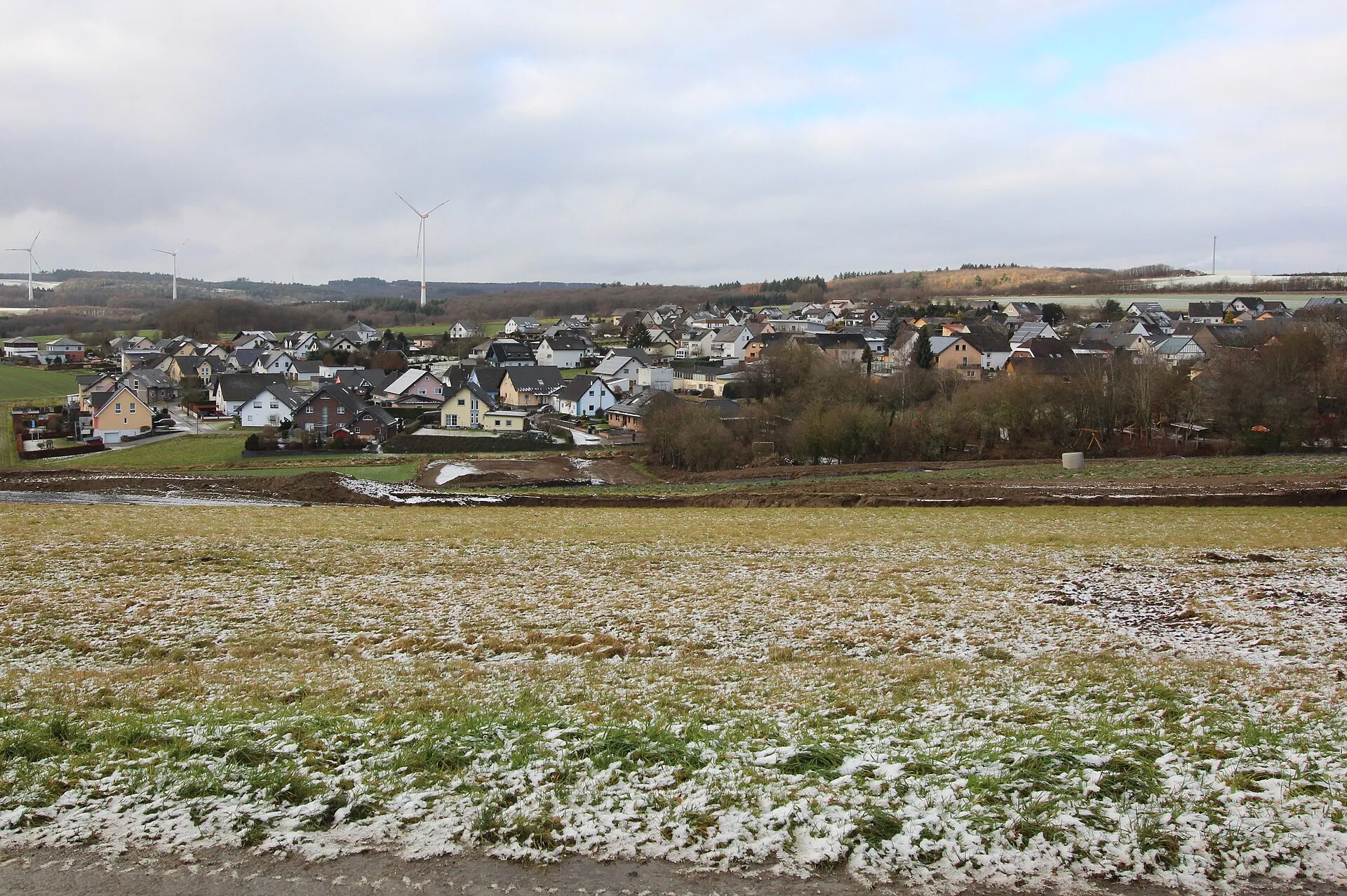 Blick über ein verschneites Feld auf die Wohnhäuser von Hambuch, dahinter bewaldete Höhen und Windräder unter grauem Himmel