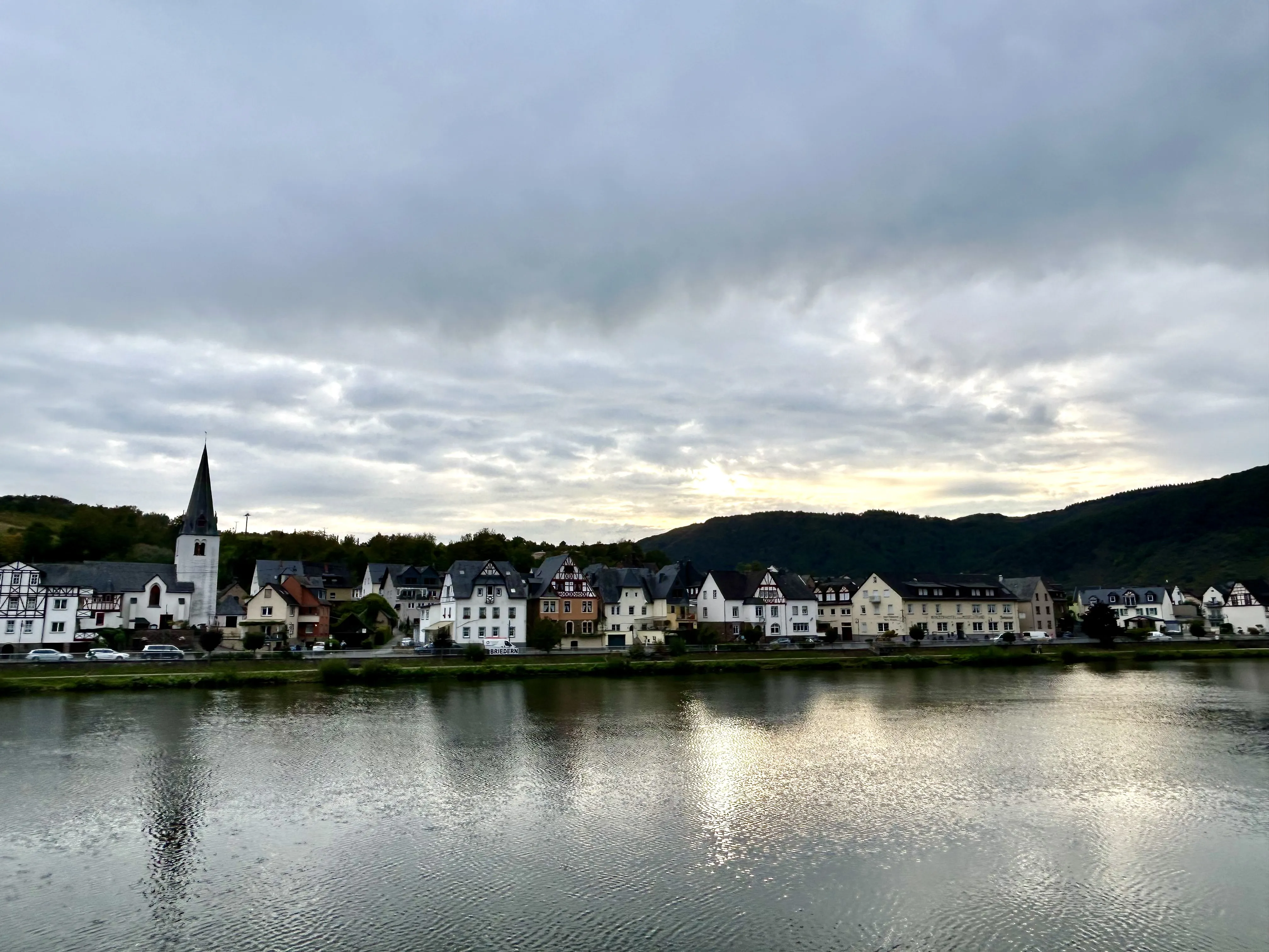Briedern von der Mosel aus: der spitze Turm der Servatiuskirche, die Häuserzeile am Ufer, dahinter bewaldete Hänge