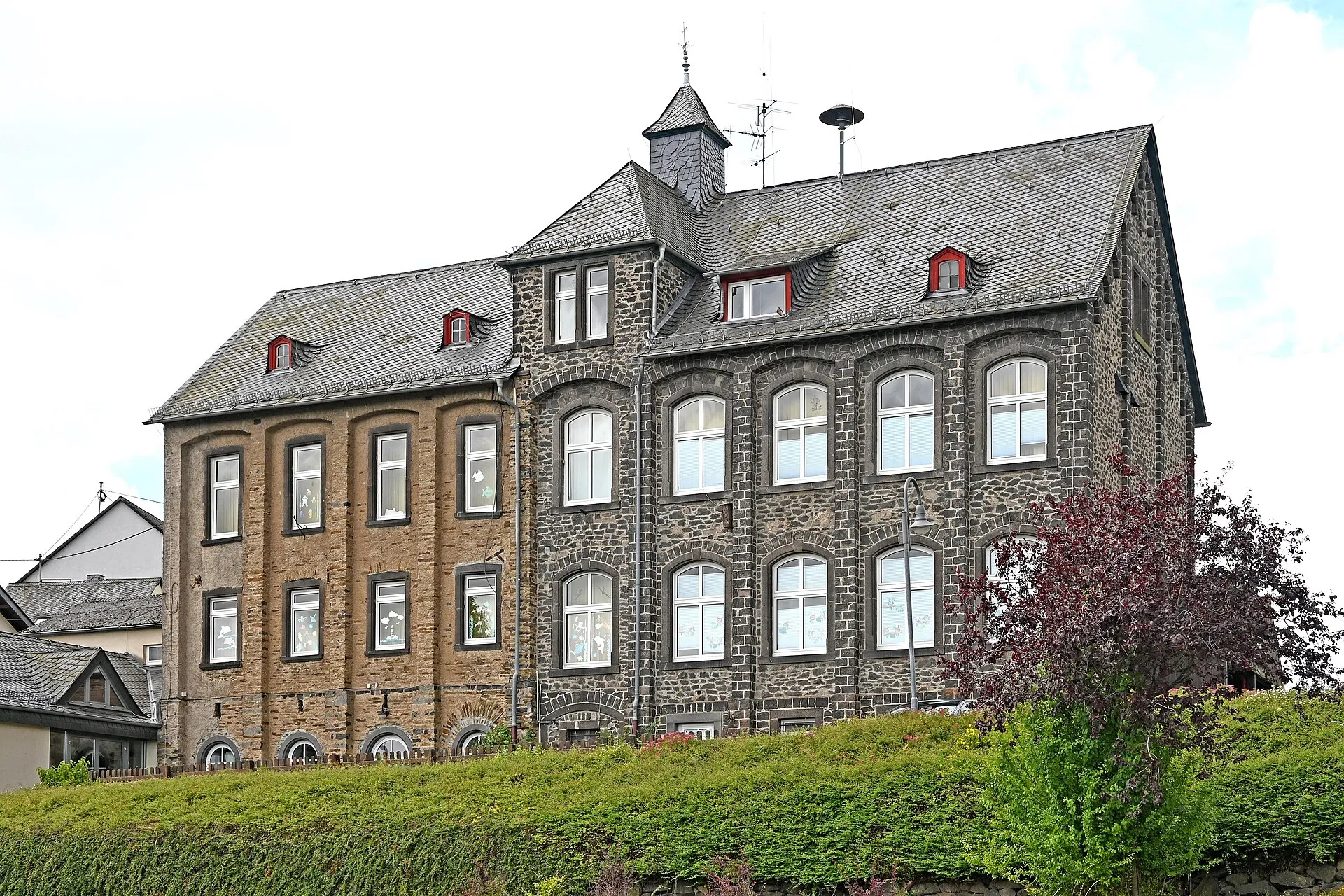 Dreigeschossiges Bruchsteingebäude mit Schieferdach und Dachreiter hinter einer hohen Hecke, an den Fenstern bunte Bilder
