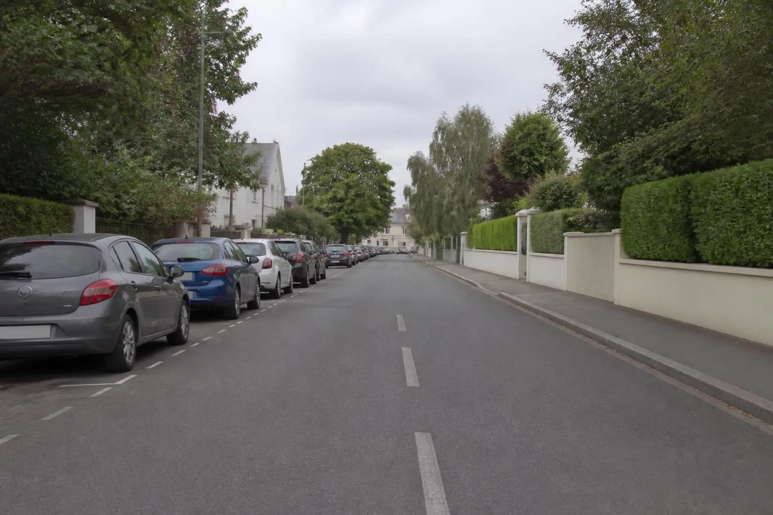 Wohnstraße im Morgenlicht, links eine Reihe parkender Autos, rechts ein freier Bordstein vor Hecke und heller Mauer