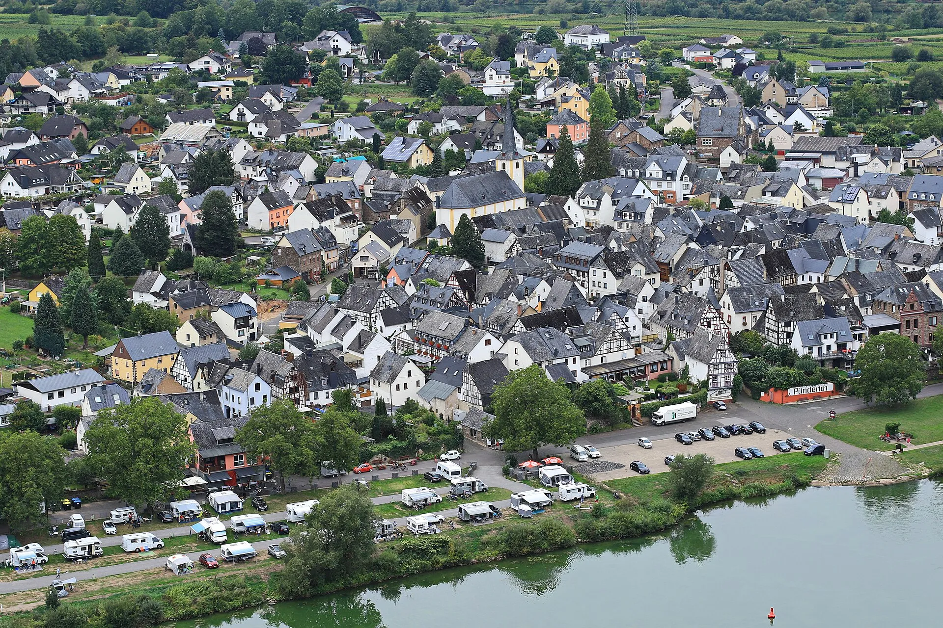 Blick von oben auf Pünderich mit Schieferdächern, Fachwerkhäusern und der Pfarrkirche, davor Wohnmobile am Moselufer